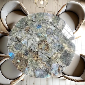 May include: A circular dining table with a polished stone surface, displaying a mosaic of grey, blue, and brown tones. The table is encircled by four upholstered chairs with light grey seats and wooden frames. A decorative light fitting hangs above.