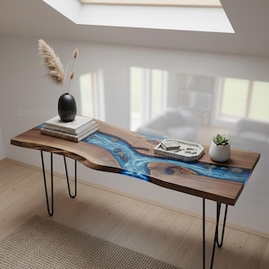 Può includere: Un tavolo in legno con un design a fiume in resina blu e gambe a forcina nere. Il tavolo presenta un bordo vivo ed è decorato con un vaso nero, libri, un piccolo vassoio e una pianta grassa in vaso. Dimensioni non visibili.