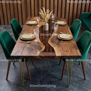 May include: A wooden dining table with a dark brown epoxy resin center, surrounded by a natural wood grain. The table is set with gold-rimmed plates, silverware, and glassware. Green velvet chairs with gold legs surround the table. A vase of dried flowers sits in the center.
