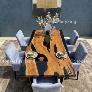 Puede incluir: Una mesa de comedor rectangular de madera con un centro de resina epoxi negra, rodeada de seis sillas tapizadas de color azul claro. La mesa está puesta con platos y cuencos, y decorada con jarrones de flores. Una pintura cuelga de la pared.