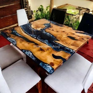 May include: A rectangular wooden table with a live edge design, featuring a dark blue epoxy resin river. The table is surrounded by white and black chairs, creating a modern dining set. The table's dimensions are approximately 60 inches long and 36 inches wide.