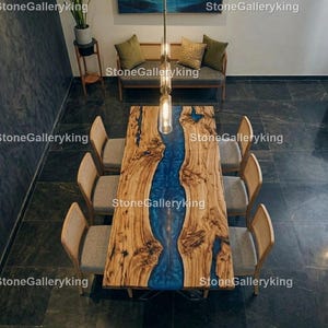 Könnte beinhalten: Ein handgefertigter Esstisch aus Holz mit einem blauen Epoxidharzfluss in der Mitte. Der Tisch ist von sechs passenden Stühlen mit geflochtenen Rückenlehnen und grau gepolsterten Sitzen umgeben. Der Tisch ist ca. 244 cm lang und 102 cm breit.