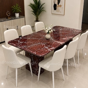 Könnte beinhalten: Ein rechteckiger Esstisch mit roter Marmorplatte und passendem Sockel. Acht weiße Stühle mit Metallbeinen umgeben den Tisch. Eine Vase mit weißen Blumen steht auf dem Tisch. Der Raum hat ein modernes Design.