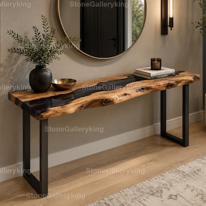 Könnte beinhalten: Eine Konsole mit Live-Edge-Design und einem schwarzen Epoxidharz-Fluss. Der Tisch hat eine natürliche Holzmaserung und steht auf schwarzen Metallbeinen. Eine schwarze Vase mit Grün, eine goldene Schale und Bücher befinden sich auf dem Tisch. Ein runder Spiegel und eine Wandleuchte sind im Hintergrund.