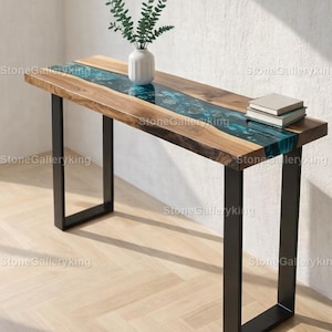 Könnte beinhalten: Ein Holztisch mit einem Flussdesign aus türkisfarbenem Harz. Der Tisch hat eine rechteckige Platte mit dunklem Holzrand und schwarzen Metallbeinen. Eine weiße Vase mit Grün und zwei Bücher stehen auf dem Tisch. Der Tisch ist ca. 152 cm lang.
