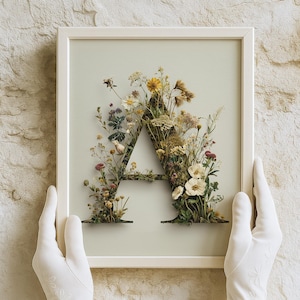 Puede incluir: Una obra de arte enmarcada con la letra "A" hecha de flores y follaje secos. El marco blanco rodea un fondo verde claro, con el arreglo floral que muestra una variedad de colores y texturas. La obra de arte está sostenida por manos enguantadas.