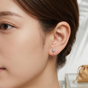 May include: A close-up of a person's ear with a small, round, diamond stud earring and a gold hoop earring. The diamond earring is surrounded by a halo of smaller diamonds. The hoop earring is a simple, thin gold band.