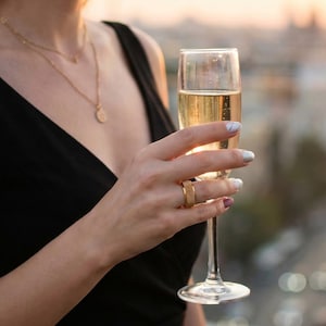 May include: A gold-colored ring with a ribbed design is worn on a finger. A champagne flute is held in the hand. The person is wearing a black dress and gold necklace. The background is blurred, with a cityscape visible.
