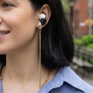 May include: A gold chain for wireless earbuds. The chain is attached to black earbud holders and dangles down. The person in the image is wearing a blue and white striped shirt.