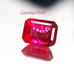May include: A vibrant, rectangular-cut red gemstone with a faceted surface that catches the light, displaying deep crimson hues. The word Gemsroot is printed in a small, dark font above the stone, which rests on a reflective surface.