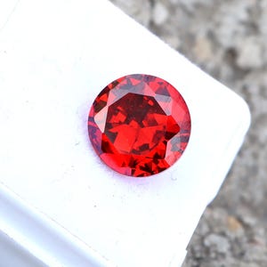 Op de afbeelding: Een gefacetteerde, ronde, helderrode edelsteen. De edelsteen staat tegen een witte achtergrond, met een grijs, getextureerd oppervlak op de achtergrond. De slijping van de edelsteen creëert meerdere facetten die licht reflecteren, waardoor hij een briljante uitstraling krijgt. Deze edelsteen kan in sieraden worden gebruikt.