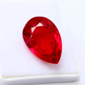 May include: A vibrant, pear-shaped, faceted red gemstone. The gem is displayed on a white surface, showcasing its brilliant cut and intense color. The gemstone's facets reflect light, creating a sparkling effect.