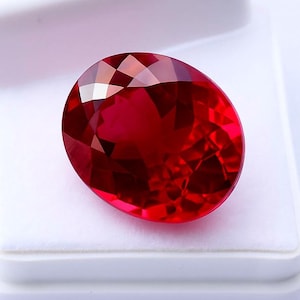 Puede incluir: Una gema roja intensa, tallada en forma ovalada. El rubí se presenta en un estuche blanco, resaltando sus múltiples facetas y su color vibrante. La piedra refleja la luz, creando un efecto brillante.