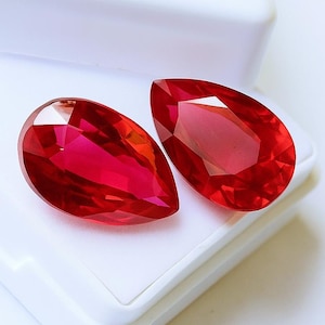 May include: Two faceted, teardrop-shaped, bright red gemstones. The gems are displayed on a white surface within a white box. The gems have a high degree of clarity and brilliance, reflecting light. The overall presentation is clean and elegant.