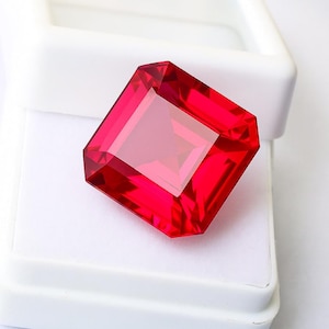 May include: A faceted, rectangular-cut, vibrant red gemstone. The gem is set against a white background inside a white box. The gem's facets reflect light, creating a brilliant display of color and clarity. The gemstone is a striking example of fine jewelry.
