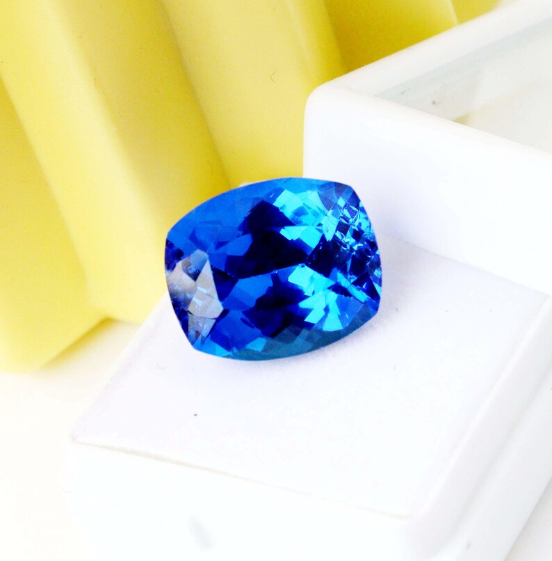K&ouml;nnte beinhalten: Ein facettierter, kissenf&ouml;rmiger Edelstein in tiefblauen Farbt&ouml;nen. Der Edelstein befindet sich vor einem wei&szlig;en Hintergrund, mit einer gelben Struktur auf der linken Seite. Die Facetten des Edelsteins reflektieren das Licht und erzeugen einen funkelnden Effekt. Geeignet f&uuml;r die Schmuckherstellung.
