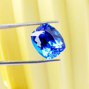 K&ouml;nnte beinhalten: Ein leuchtender, kissenf&ouml;rmig geschliffener blauer Edelstein wird von einer silbernen Pinzette vor einem gelben Hintergrund gehalten. Die Facetten des Edelsteins reflektieren das Licht und erzeugen einen funkelnden Effekt. Der Edelstein hat eine tiefe, satte blaue Farbe.