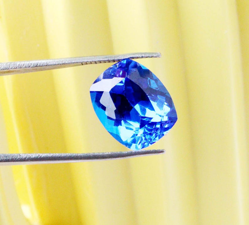 K&ouml;nnte beinhalten: Ein leuchtender, kissenf&ouml;rmig geschliffener blauer Edelstein wird von einer silbernen Pinzette vor einem gelben Hintergrund gehalten. Die Facetten des Edelsteins reflektieren das Licht und erzeugen einen funkelnden Effekt. Der Edelstein hat eine tiefe, satte blaue Farbe.
