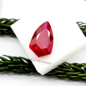 May include: A faceted, pear-shaped, vibrant red gemstone. The gem is displayed on a white surface with the text "Gemsroot" visible. Green foliage frames the gem, enhancing its color and brilliance.