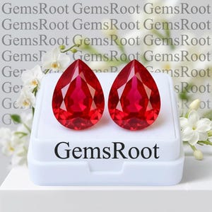 May include: Two pear-shaped, faceted red gemstones displayed on a white rectangular stand. The stand features the text GemsRoot printed in black. The stones exhibit a deep, vibrant red color with multiple triangular facets reflecting light.