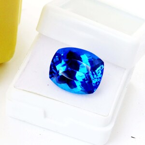 K&ouml;nnte beinhalten: Ein facettierter, kissenf&ouml;rmiger, leuchtend blauer Edelstein liegt in einer offenen wei&szlig;en Kunststoffbox. Die Facetten des Edelsteins reflektieren das Licht und erzeugen einen funkelnden Effekt. Die Box zeigt die lebendige Farbe und Form des Edelsteins.