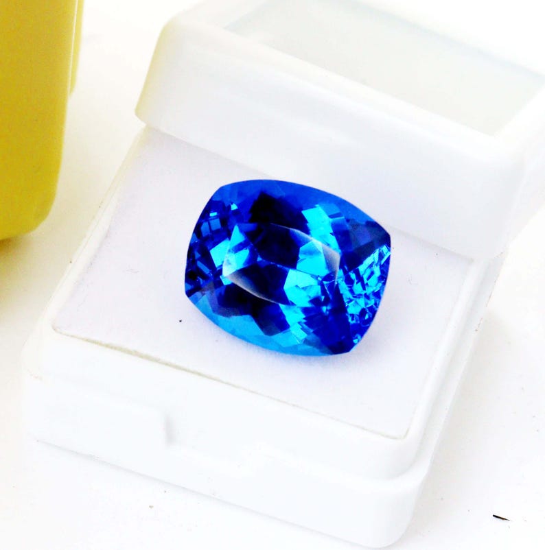 K&ouml;nnte beinhalten: Ein facettierter, kissenf&ouml;rmiger, leuchtend blauer Edelstein liegt in einer offenen wei&szlig;en Kunststoffbox. Die Facetten des Edelsteins reflektieren das Licht und erzeugen einen funkelnden Effekt. Die Box zeigt die lebendige Farbe und Form des Edelsteins.