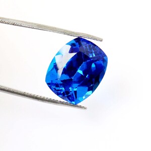 K&ouml;nnte beinhalten: Ein leuchtend blauer, kissenf&ouml;rmig geschliffener Edelstein, der von einer Pinzette vor einem wei&szlig;en Hintergrund gehalten wird. Die Facetten des Edelsteins reflektieren das Licht und erzeugen ein brillantes Farb- und Funkelspiel. Der Edelstein ist ein tiefes, sattes Blau.