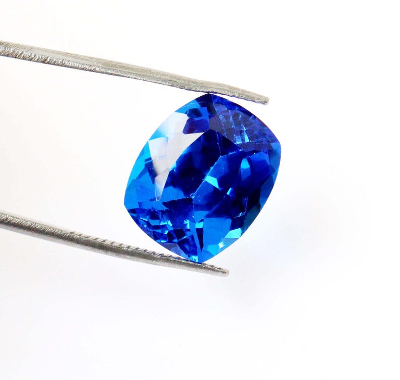 K&ouml;nnte beinhalten: Ein leuchtend blauer, kissenf&ouml;rmig geschliffener Edelstein, der von einer Pinzette vor einem wei&szlig;en Hintergrund gehalten wird. Die Facetten des Edelsteins reflektieren das Licht und erzeugen ein brillantes Farb- und Funkelspiel. Der Edelstein ist ein tiefes, sattes Blau.