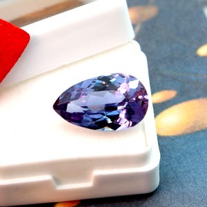 May include: A pear-shaped, faceted gemstone with a blend of purple and blue hues. The gem is set against a white background inside a white box, with a red accent visible. The stone's facets reflect light, creating a sparkling effect.