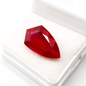 Könnte beinhalten: Ein facettierter, tiefroter Edelstein mit einer einzigartigen, länglichen Form. Der Edelstein wird in einer weißen Schachtel präsentiert, die seinen Brillantschliff und seine lebendige Farbe hervorhebt. Die Facetten des Edelsteins reflektieren das Licht.