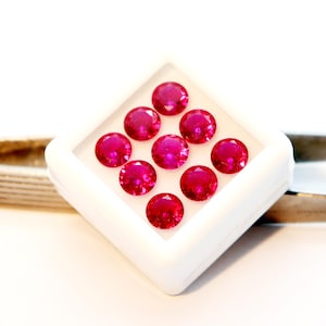 May include: Nine round, bright red gemstones are presented in a square, white container. The gemstones are evenly spaced and appear to be faceted, reflecting light. The setting is simple, highlighting the gems' colour and clarity.