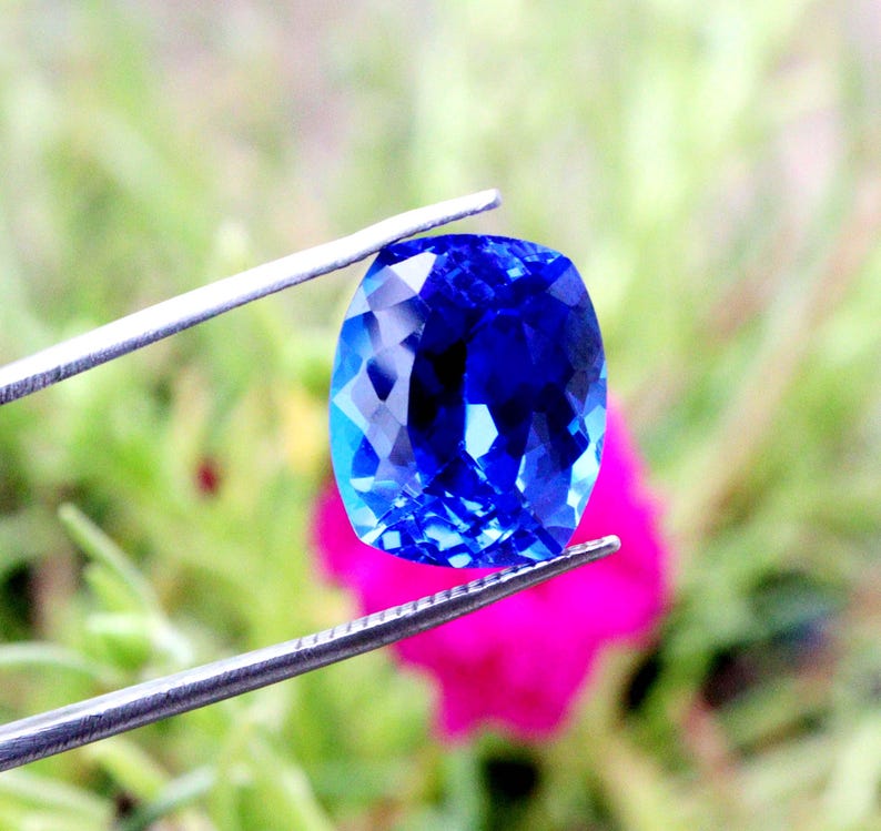 K&ouml;nnte beinhalten: Ein facettierter, kissenf&ouml;rmiger, tiefblauer Edelstein wird von einer silbernen Pinzette gehalten. Die Facetten des Edelsteins reflektieren das Licht und erzeugen ein brillantes Funkeln. Der Hintergrund zeigt verschwommenes gr&uuml;nes Laub und eine leuchtend rosa Blume.