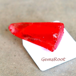 May include: A vibrant red gemstone, cut into a triangular shape, rests on a white tag. The gemstone's surface is smooth and reflects light, showcasing its rich color. The tag has the text "GemsRoot" printed on it.