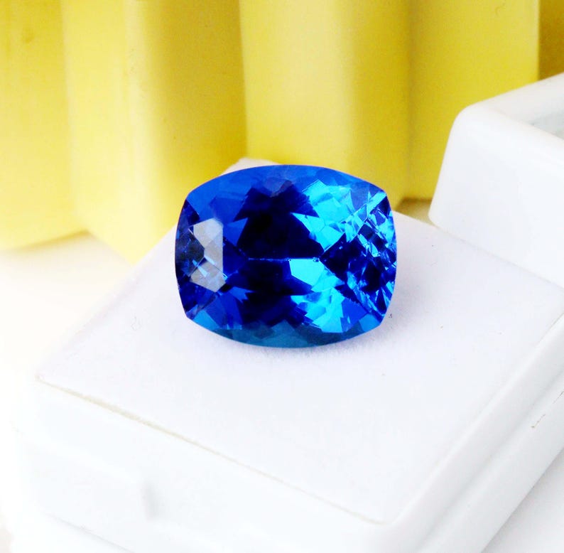 K&ouml;nnte beinhalten: Ein lebendiger, kissenf&ouml;rmig geschliffener blauer Edelstein mit Facetten, die das Licht reflektieren. Der Edelstein liegt auf einer wei&szlig;en Oberfl&auml;che mit gelbem Hintergrund. Die Farbe des Edelsteins reicht von tiefem Indigo bis zu hellem T&uuml;rkis.
