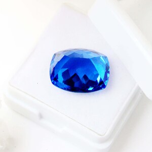K&ouml;nnte beinhalten: Ein facettierter, kissenf&ouml;rmiger, saphirblauer Edelstein. Der Edelstein befindet sich in einer wei&szlig;en Schachtel, deren Deckel teilweise ge&ouml;ffnet ist. Die Facetten des Edelsteins reflektieren das Licht und erzeugen ein brillantes Farb- und Glitzerspiel. Der Edelstein ist ein tiefes, sattes Blau.