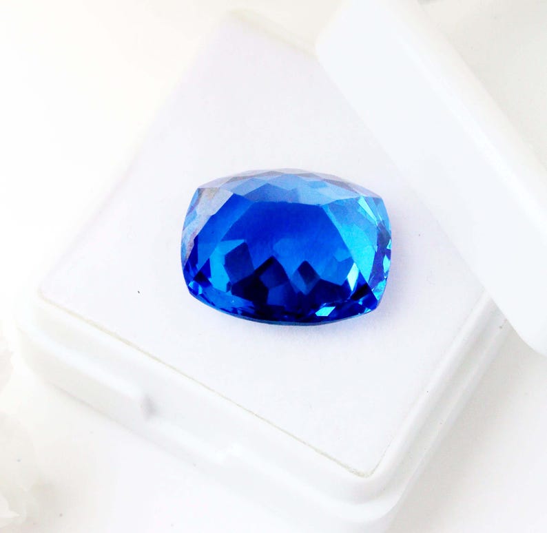 K&ouml;nnte beinhalten: Ein facettierter, kissenf&ouml;rmiger, saphirblauer Edelstein. Der Edelstein befindet sich in einer wei&szlig;en Schachtel, deren Deckel teilweise ge&ouml;ffnet ist. Die Facetten des Edelsteins reflektieren das Licht und erzeugen ein brillantes Farb- und Glitzerspiel. Der Edelstein ist ein tiefes, sattes Blau.