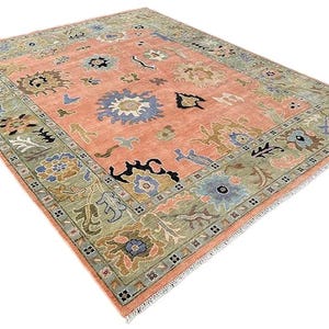 以下が含まれることがあります： 中央がコーラル色で、縁がセージグリーンの長方形のラグ。青、茶色、黒の色合いで花柄と幾何学模様が施されています。ラグの端はフリンジ加工が施されています。