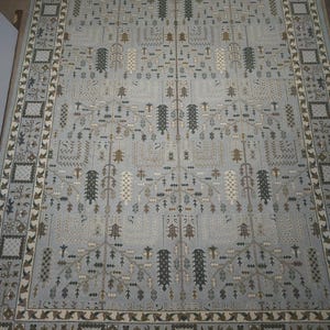 Puede incluir: Una alfombra rectangular grande con un fondo azul claro y un patrón intrincado. El diseño presenta motivos repetidos de árboles estilizados, hojas y formas geométricas en tonos de marrón, beige y verde oscuro. La alfombra tiene un borde detallado.