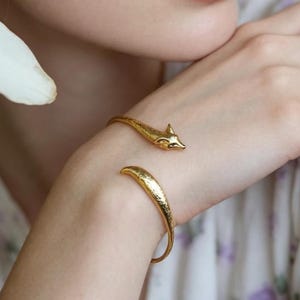 May include: A gold-toned open cuff bracelet featuring a sculpted fox head at one end and a tapered tail at the other, worn on the wrist. The metal surface has a textured finish, and the design is positioned near yellow and white flowers.