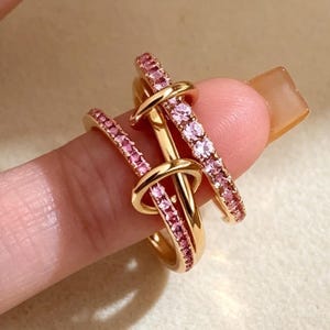 May include: A gold-toned ring featuring a unique interlocking design with two bands. One band is plain polished gold, whilst the other is encrusted with small, sparkling pink stones. The two bands are joined together in a knot-like structure.