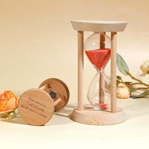 May include: A wooden hourglass with red sand inside a clear glass bulb. The hourglass is supported by a wooden frame. The text "If you look at me, I will melt gently like the snow on a volcano" is engraved on the base.