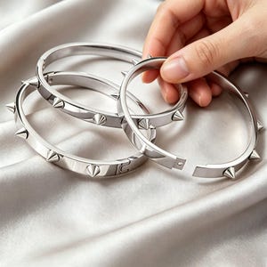 以下が含まれることがあります： 光沢のある仕上げと等間隔に配置された円錐形のスパイクが特徴の、シルバー調の金属製バングルブレスレット3点。各円形のブレスレットにはヒンジ式の留め具が付いており、手首に合わせて安全に開閉できるようになっています。