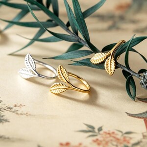 May include: Two open-style rings shaped like twin leaves, one finished in polished silver and the other in polished gold. Each ring features a textured leaf design that curves around the finger, with one leaf pointing upward and one downward.