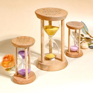 Può includere: Tre clessidre in legno con bulbi di vetro trasparente e sabbia colorata. I colori della sabbia sono giallo, viola e rosa. Ogni clessidra ha una base e una parte superiore in legno con testo inciso.