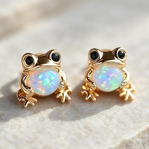 May include: A pair of gold-toned stud earrings shaped like frogs. Each frog features a round, iridescent white opal-like stone as its belly and two small, black circular eyes. The metal has a polished finish, and the frogs are depicted in a seated position.