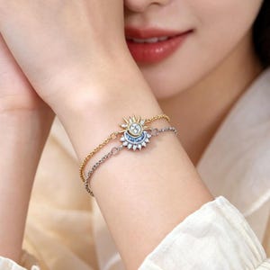 May include: A dual-strand bracelet featuring a gold chain with a sun charm and a silver chain with a crescent moon charm. The sun charm has a central clear stone, and the moon charm is accented with small blue and clear stones.