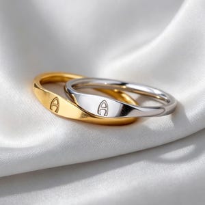 May include: Two thin metal rings, one gold-toned and one silver-toned, each featuring a flat, polished surface section engraved with the capital letter A. The rings are interlocked, showcasing a smooth, rounded band design with a metallic finish.