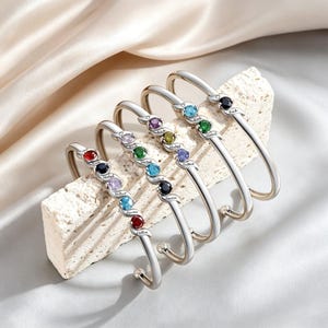 May include: Five silver-toned bangles with colorful gemstones. Each bangle features a series of gemstones in red, black, purple, blue, green, yellow, and black. The gemstones are set in a row, and each bangle has a name engraved on it.