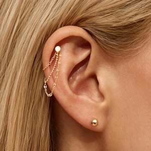 May include: A gold-toned ear cuff featuring a small circular stud connected to three delicate dangling chains that loop back to the stud. A separate, matching gold-toned spherical ball stud earring is positioned in the lower earlobe.