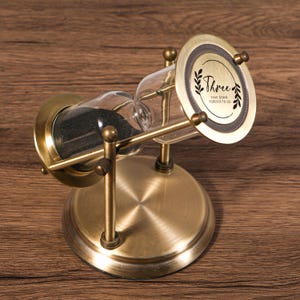 Könnte beinhalten: Eine Sanduhr im antiken Stil mit Bronze-Finish. Die Glaskammer enthält schwarzen Sand. Die obere runde Platte trägt das Wort "Three" und den Satz "Year Down Forever to Go". Die Sanduhr steht auf einem bronzefarbenen Sockel.